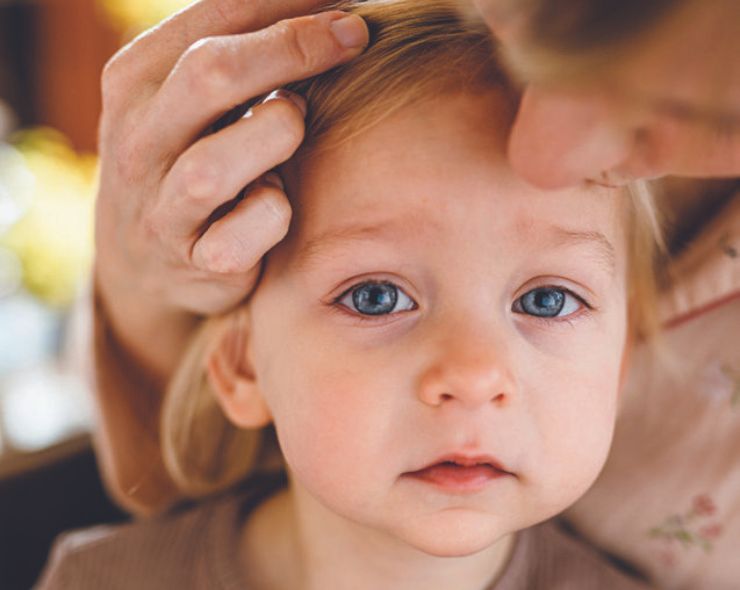 A mother holds her child's face as she places a kiss on her forehead. The child has blonde hair and blue eyes. She looks unsettled.