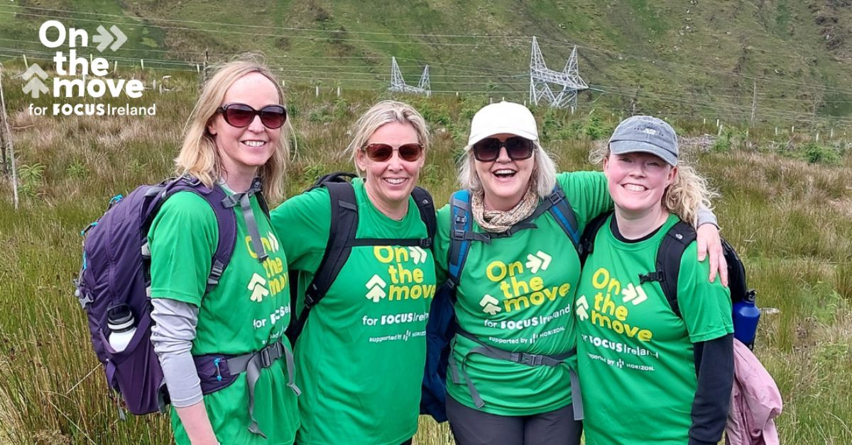 Four Focus Ireland hikers stand arm in arm smileing to camera. They are all wearing Green On The Move t-shirts.